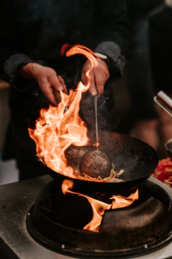 Wok z płomieniami, chochlą i smażonym makaronem.